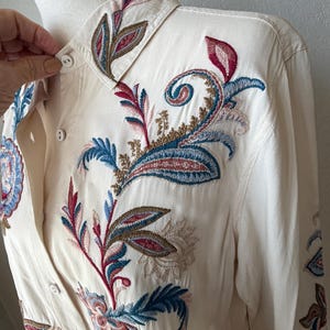 May include: Cream-colored button-down shirt with intricate floral embroidery in shades of red, blue, and brown. The embroidery is detailed on the collar, front, and sleeves. The shirt has long sleeves and a classic collar.