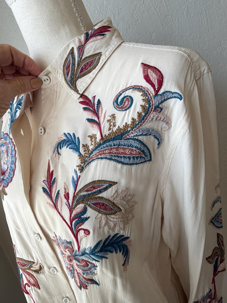 May include: Cream-colored button-down shirt with intricate floral embroidery in shades of red, blue, and brown. The embroidery is detailed on the collar, front, and sleeves. The shirt has long sleeves and a classic collar.