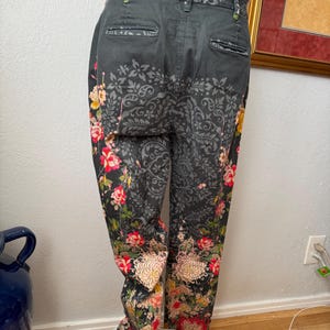 Puede incluir: Pantalones gris carb&oacute;n oscuro con un patr&oacute;n de damasco negro en la parte superior y estampados florales vibrantes en tonos rojo, rosa, amarillo y verde en la parte inferior de las piernas, con dos bolsillos traseros y cinturilla.