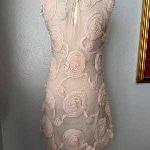 May include: A sleeveless, beige dress adorned with raised, circular floral appliqu&eacute;s. The dress features a keyhole opening at the back and is displayed on a mannequin. The dress is a light, airy fabric, perfect for a special occasion.