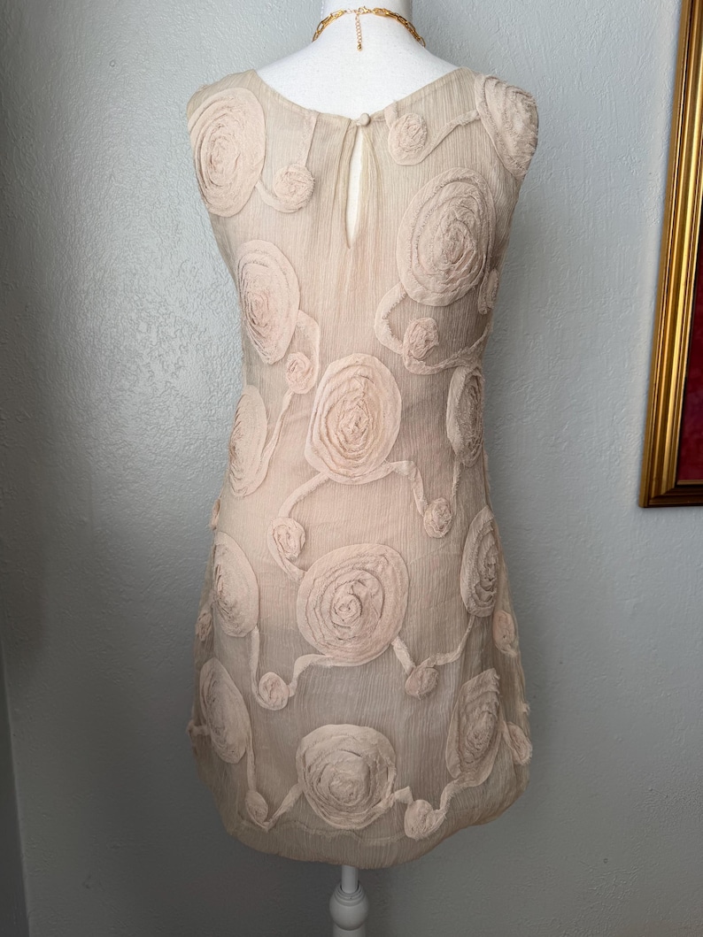 May include: A sleeveless, beige dress adorned with raised, circular floral appliqu&eacute;s. The dress features a keyhole opening at the back and is displayed on a mannequin. The dress is a light, airy fabric, perfect for a special occasion.