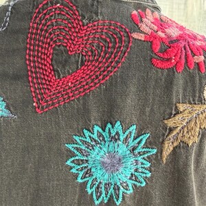 以下が含まれることがあります： カラフルな刺繍の花柄とハートのデザインが施された、濃いグレーのデニムジャケットのクローズアップ。刺繍は赤、ピンク、青、茶色の色合いで、ジャケットに鮮やかなタッチを加えています。