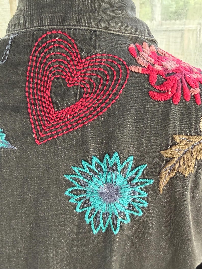 以下が含まれることがあります： カラフルな刺繍の花柄とハートのデザインが施された、濃いグレーのデニムジャケットのクローズアップ。刺繍は赤、ピンク、青、茶色の色合いで、ジャケットに鮮やかなタッチを加えています。