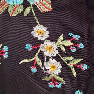 May include: Close-up of black fabric with colorful floral embroidery. The embroidery features white, yellow, blue, and red flowers and green leaves. The stitching is detailed and creates a textured effect on the dark background.