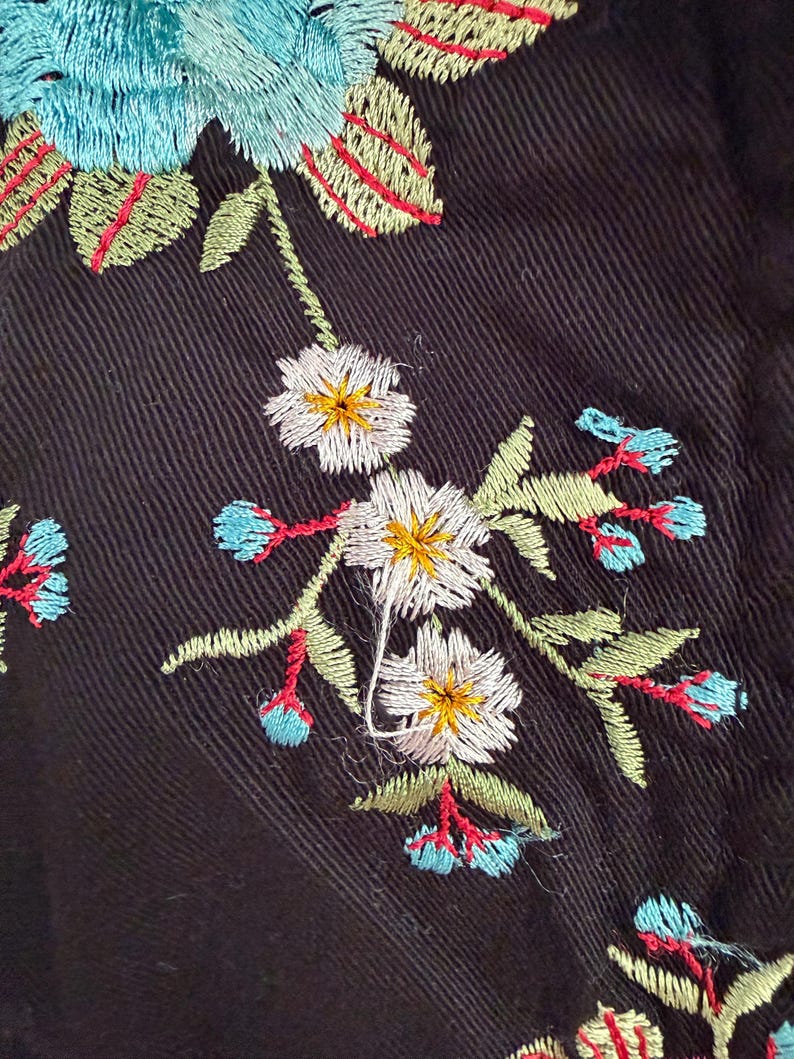 May include: Close-up of black fabric with colorful floral embroidery. The embroidery features white, yellow, blue, and red flowers and green leaves. The stitching is detailed and creates a textured effect on the dark background.