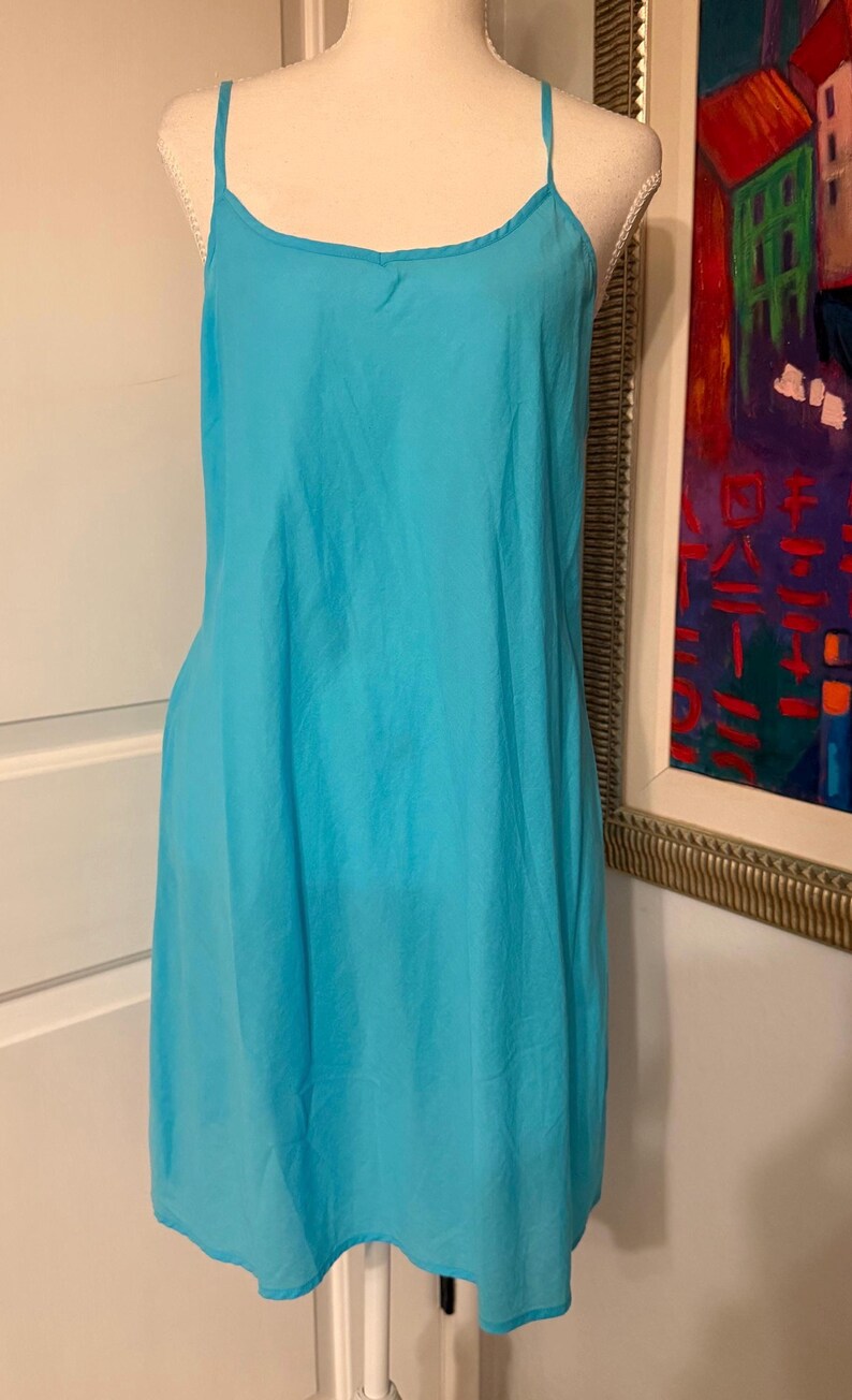 Puede incluir: Un vestido lencero turquesa con finas tiras de espagueti. El vestido se exhibe en un maniqu&iacute;. La tela parece ligera y tiene un ligero brillo. El vestido llega hasta la rodilla.