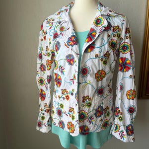 Puede incluir: Chaqueta blanca con bordado floral con flores rojas, amarillas, azules y verdes. La chaqueta tiene cuello y cierre de botones. Se ve una camiseta azul claro debajo.