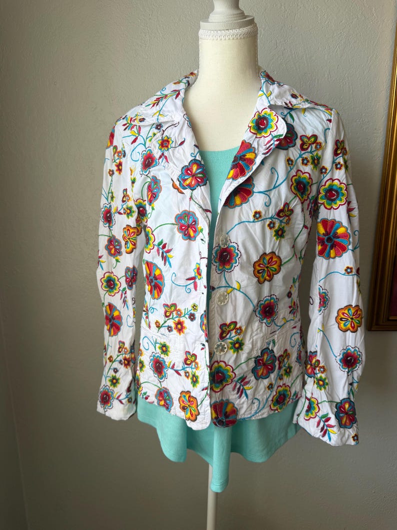 Puede incluir: Chaqueta blanca con bordado floral con flores rojas, amarillas, azules y verdes. La chaqueta tiene cuello y cierre de botones. Se ve una camiseta azul claro debajo.