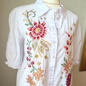 May include: White button-down shirt with colorful floral embroidery. The embroidery features red, pink, green, and gold thread in a detailed floral pattern. The shirt has short sleeves and a collared neckline.