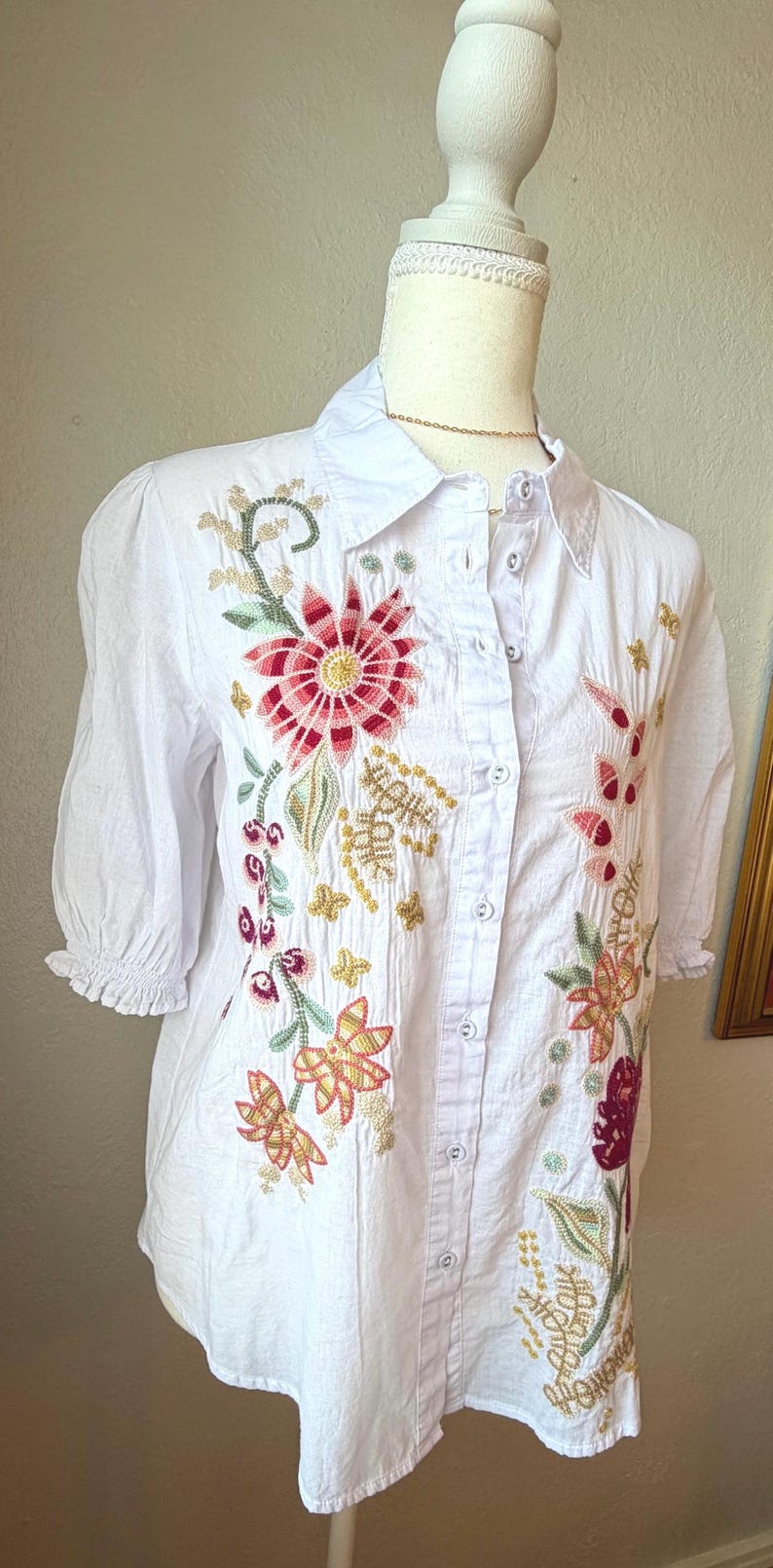 May include: White button-down shirt with colorful floral embroidery. The embroidery features red, pink, green, and gold thread in a detailed floral pattern. The shirt has short sleeves and a collared neckline.
