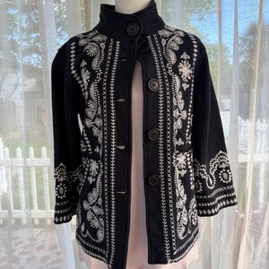 K&ouml;nnte beinhalten: Eine schwarze Jacke mit aufwendiger wei&szlig;er Stickerei. Die Jacke hat einen hohen Kragen, Knopfverschl&uuml;sse und Dreiviertel&auml;rmel. Die Stickerei umfasst Blumen- und Paisley-Muster, die einen detaillierten und eleganten Look erzeugen. Die Jacke wird auf einer Schaufensterpuppe pr&auml;sentiert.