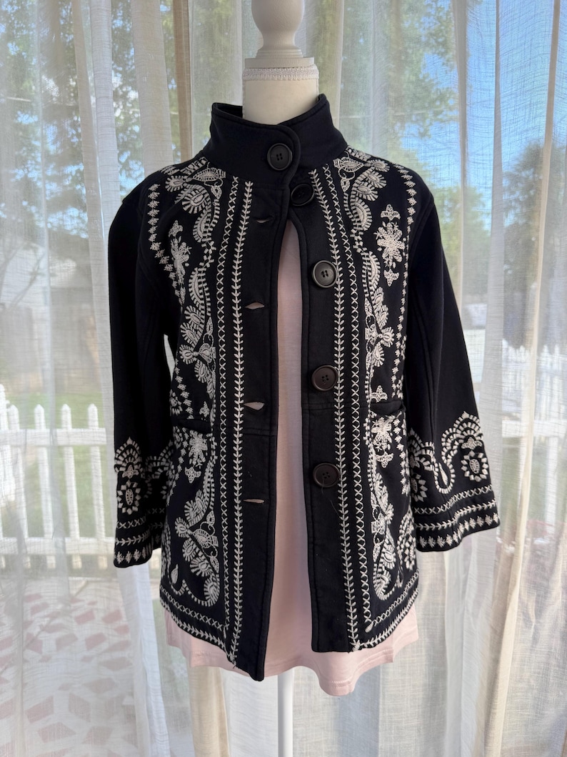 K&ouml;nnte beinhalten: Eine schwarze Jacke mit aufwendiger wei&szlig;er Stickerei. Die Jacke hat einen hohen Kragen, Knopfverschl&uuml;sse und Dreiviertel&auml;rmel. Die Stickerei umfasst Blumen- und Paisley-Muster, die einen detaillierten und eleganten Look erzeugen. Die Jacke wird auf einer Schaufensterpuppe pr&auml;sentiert.