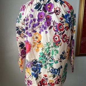 May include: A cream-colored blouse with a vibrant floral pattern featuring purple, red, blue, and orange flowers. The long-sleeved top has a round neckline and a gathered waist detail, creating a feminine silhouette.