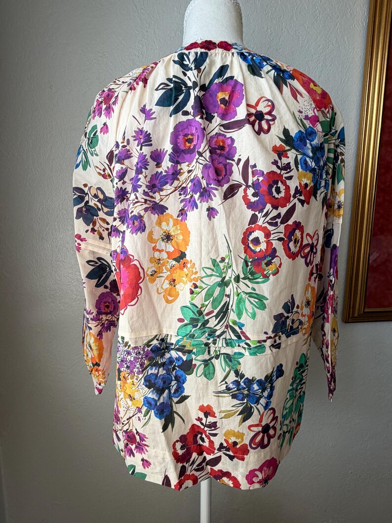 May include: A cream-colored blouse with a vibrant floral pattern featuring purple, red, blue, and orange flowers. The long-sleeved top has a round neckline and a gathered waist detail, creating a feminine silhouette.