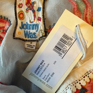 May include: Close-up of a white dress with colorful embroidered floral and geometric patterns. A tag with the brand name "Johnny Was" and product information is attached. The dress has a small size.