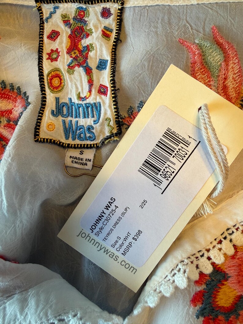 May include: Close-up of a white dress with colorful embroidered floral and geometric patterns. A tag with the brand name "Johnny Was" and product information is attached. The dress has a small size.
