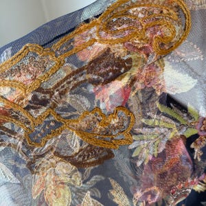 May include: Close-up of a sheer fabric with a floral pattern in shades of pink, brown, and green. The fabric is overlaid with a dark blue mesh and embellished with gold-colored embroidery.