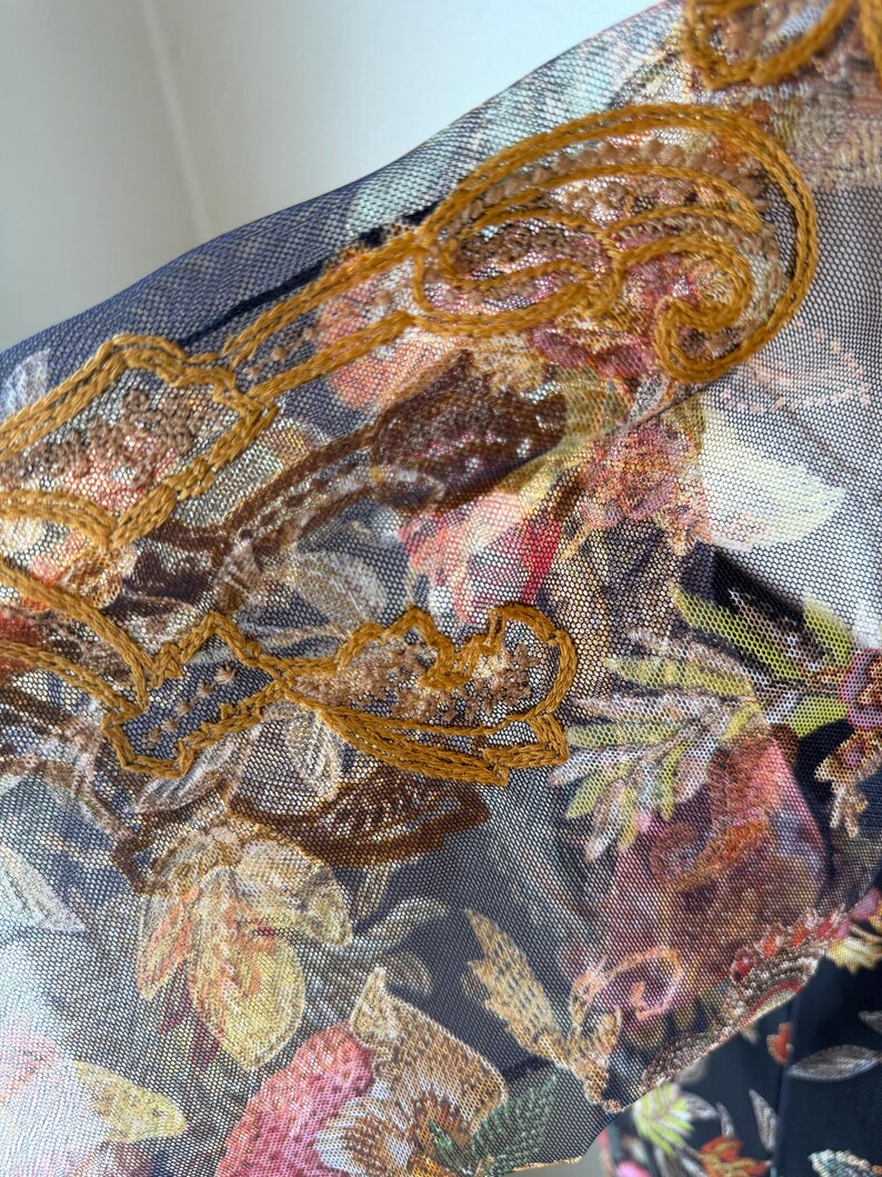 May include: Close-up of a sheer fabric with a floral pattern in shades of pink, brown, and green. The fabric is overlaid with a dark blue mesh and embellished with gold-colored embroidery.