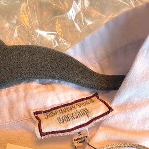 May include: Close-up of a white shirt with a collar and a label that reads "JOHNNY WAS WORKSHOP". The shirt is hanging on a gray hanger. The label indicates the shirt is size small and made in China.