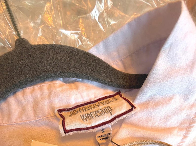 May include: Close-up of a white shirt with a collar and a label that reads "JOHNNY WAS WORKSHOP". The shirt is hanging on a gray hanger. The label indicates the shirt is size small and made in China.