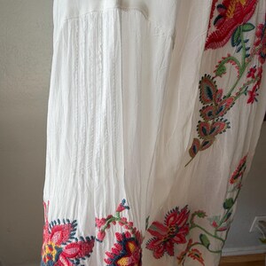 May include: White dress with colorful floral embroidery. The embroidery features red, pink, yellow, and blue flowers with green leaves. The dress has pleats and a flowing design.