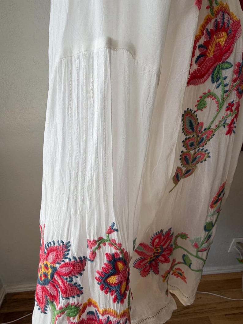 May include: White dress with colorful floral embroidery. The embroidery features red, pink, yellow, and blue flowers with green leaves. The dress has pleats and a flowing design.