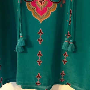 May include: Teal-colored tunic with a detailed floral and geometric design in pink, gold, and black. The tunic features a tassel tie neckline and decorative tassels. The fabric appears lightweight and flowing.