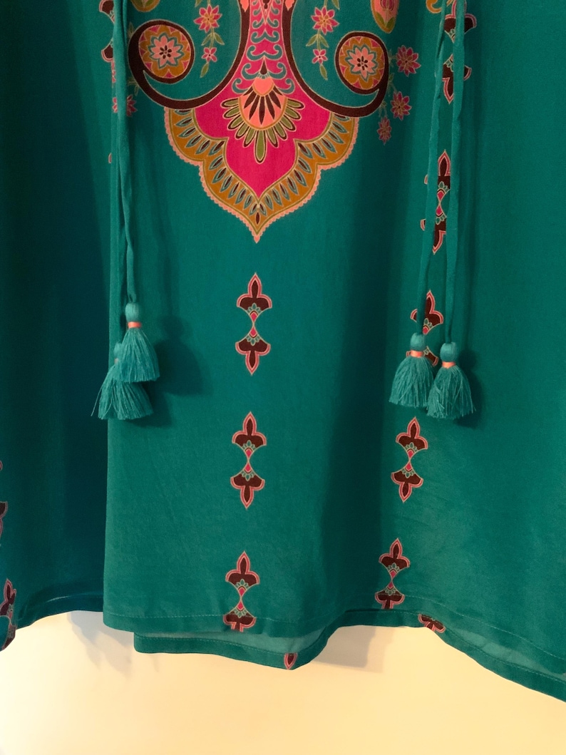 May include: Teal-colored tunic with a detailed floral and geometric design in pink, gold, and black. The tunic features a tassel tie neckline and decorative tassels. The fabric appears lightweight and flowing.