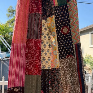 Peut inclure: Une longue jupe patchwork avec une variété de motifs de tissus colorés. La jupe est composée de différents morceaux de tissu rectangulaires cousus ensemble. Les couleurs incluent le rouge, le rose, le noir, le vert et le jaune. La jupe est suspendue à une corde à linge.