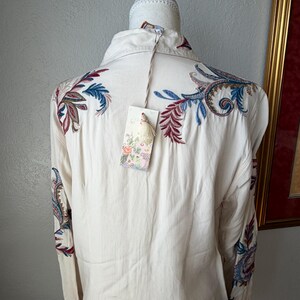 May include: Cream-colored button-down shirt with intricate embroidered floral and paisley designs in shades of blue, red, and green. The long-sleeved shirt has a tag attached. The shirt is displayed on a mannequin.