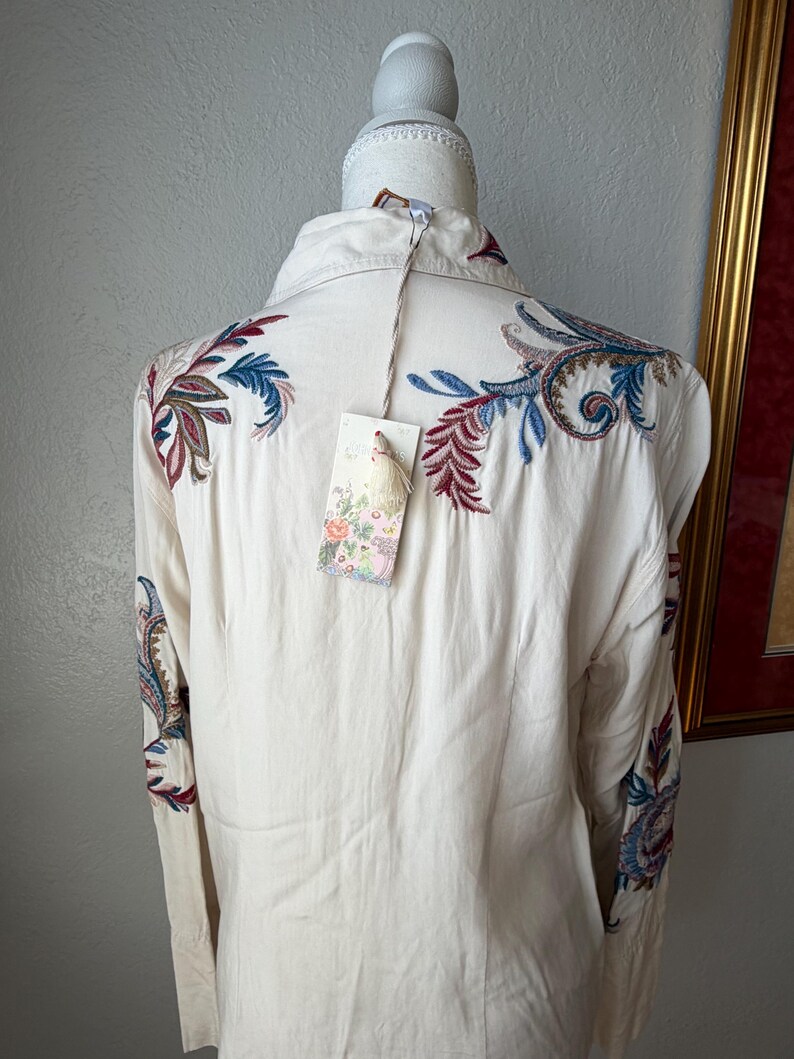 May include: Cream-colored button-down shirt with intricate embroidered floral and paisley designs in shades of blue, red, and green. The long-sleeved shirt has a tag attached. The shirt is displayed on a mannequin.