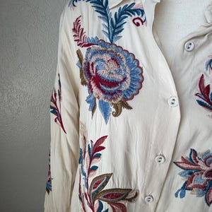 May include: Cream-colored button-down shirt with intricate floral embroidery in shades of blue, red, and green. The long-sleeved shirt features a classic collar and button closures, showcasing detailed stitching.