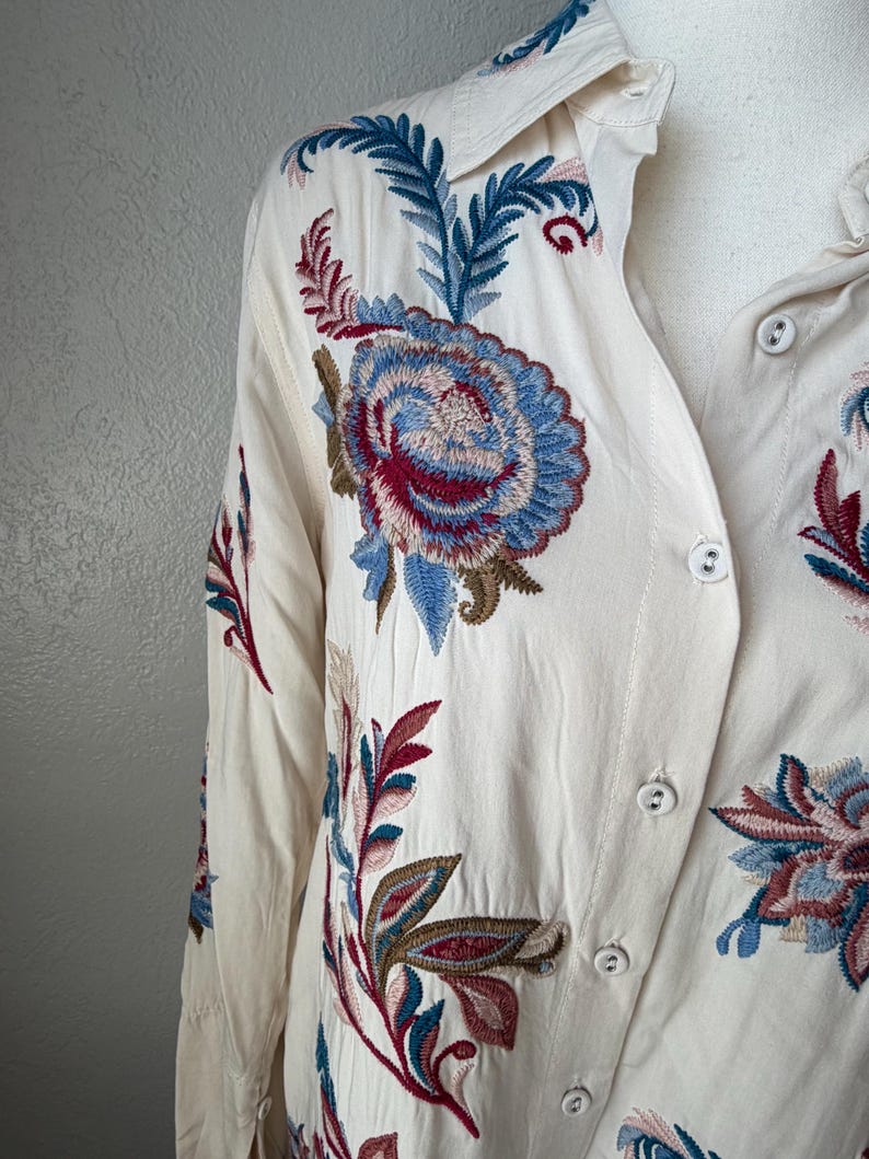May include: Cream-colored button-down shirt with intricate floral embroidery in shades of blue, red, and green. The long-sleeved shirt features a classic collar and button closures, showcasing detailed stitching.