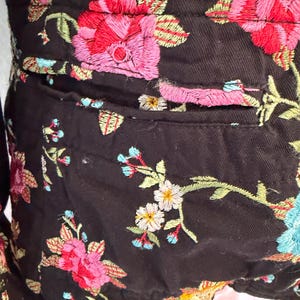 May include: Black jacket with colorful floral embroidery. The embroidery features pink, red, and blue flowers with green leaves. The jacket has a pocket and is made of a dark fabric.