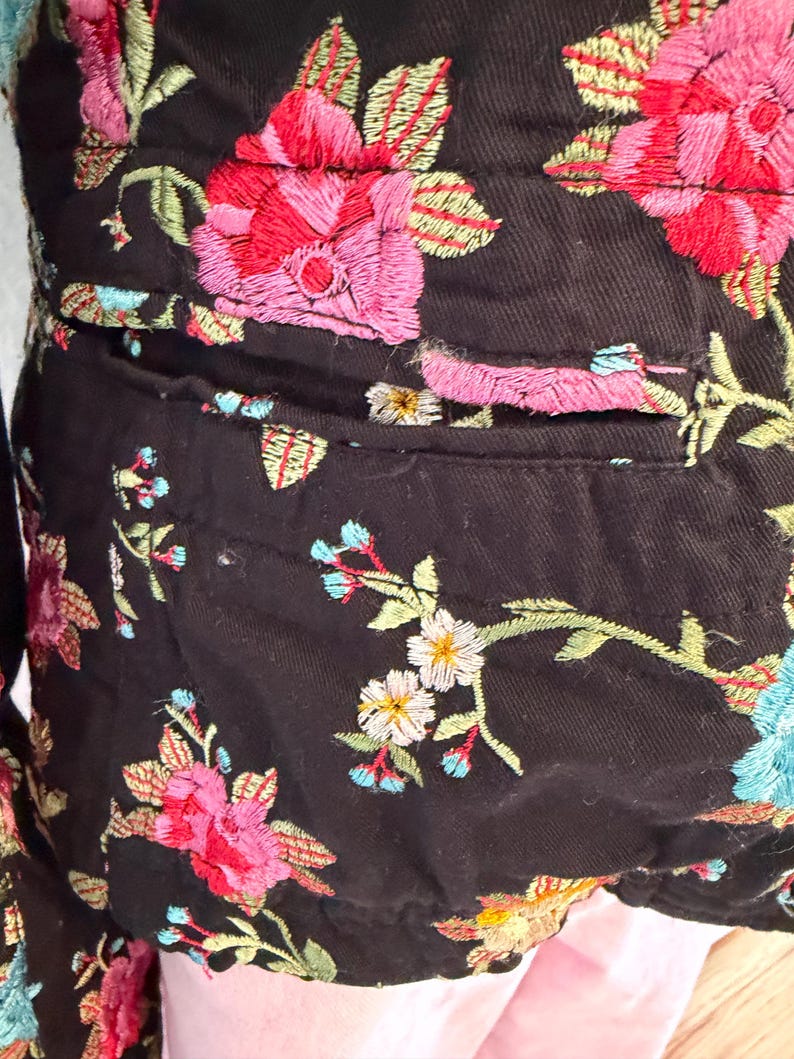May include: Black jacket with colorful floral embroidery. The embroidery features pink, red, and blue flowers with green leaves. The jacket has a pocket and is made of a dark fabric.