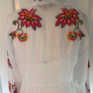 May include: White dress with colorful floral embroidery on the back and sleeves. The embroidery features red, pink, yellow, and blue flowers with green stems and leaves. The dress has a high neckline and long sleeves.