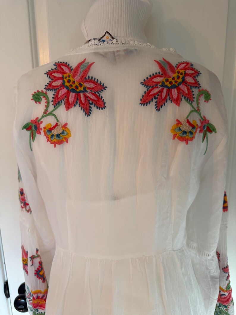 May include: White dress with colorful floral embroidery on the back and sleeves. The embroidery features red, pink, yellow, and blue flowers with green stems and leaves. The dress has a high neckline and long sleeves.