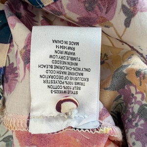 May include: Close-up of a floral print fabric with a white tag attached. The tag displays care instructions and fabric composition: 100% cotton, 100% polyester. The fabric features a mix of pink, purple, and green floral patterns.