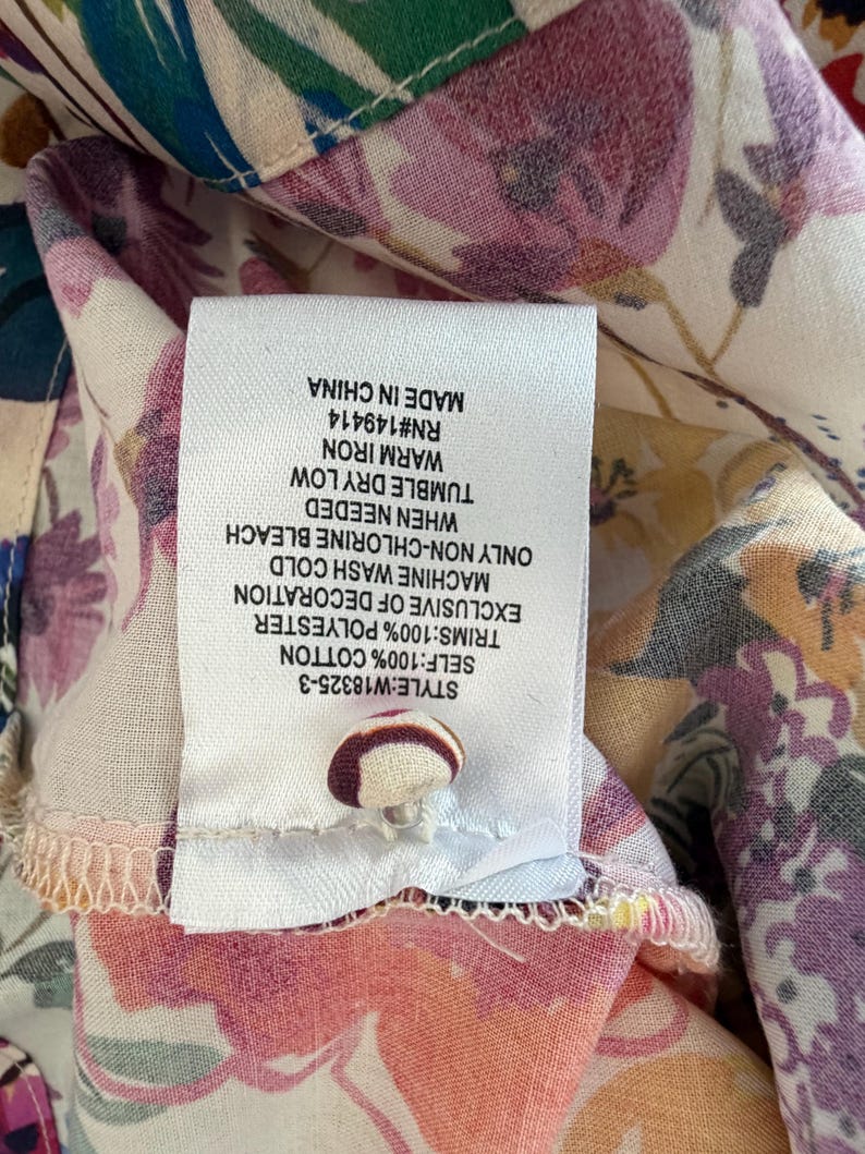 May include: Close-up of a floral print fabric with a white tag attached. The tag displays care instructions and fabric composition: 100% cotton, 100% polyester. The fabric features a mix of pink, purple, and green floral patterns.