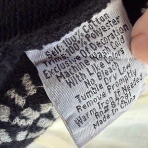 K&ouml;nnte beinhalten: Nahaufnahme eines Kleidungsetiketts mit Pflegehinweisen. Das Etikett ist grau mit schwarzem Text, und der Stoff hat ein schwarz-wei&szlig;es Muster. Auf dem Etikett steht "Self: 100% Cotton, Trims: 100% Polyester, Machine Wash Cold."