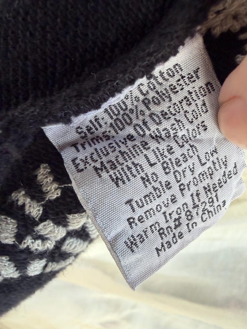 K&ouml;nnte beinhalten: Nahaufnahme eines Kleidungsetiketts mit Pflegehinweisen. Das Etikett ist grau mit schwarzem Text, und der Stoff hat ein schwarz-wei&szlig;es Muster. Auf dem Etikett steht "Self: 100% Cotton, Trims: 100% Polyester, Machine Wash Cold."