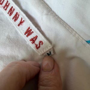 Puede incluir: Primer plano de una etiqueta de tela blanca con el texto rojo "JOHNNY WAS". La etiqueta est&aacute; adherida a una prenda blanca. Se ve una peque&ntilde;a etiqueta con la letra "M".