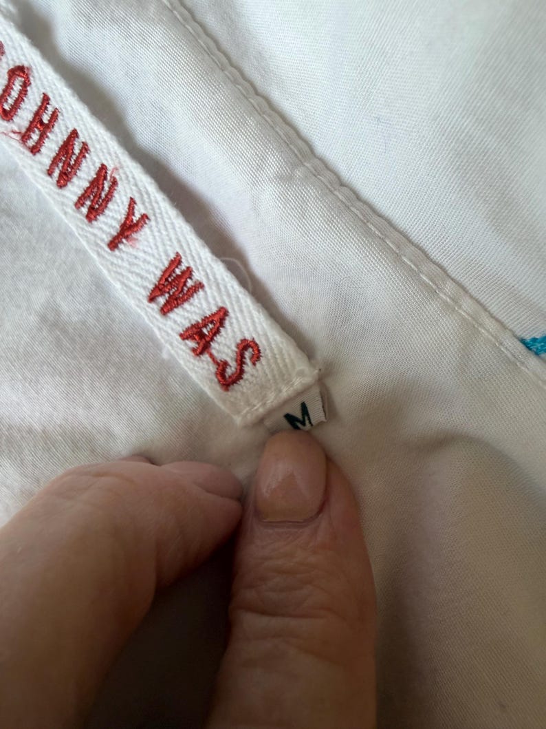 Puede incluir: Primer plano de una etiqueta de tela blanca con el texto rojo "JOHNNY WAS". La etiqueta est&aacute; adherida a una prenda blanca. Se ve una peque&ntilde;a etiqueta con la letra "M".