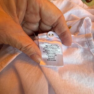 May include: Close-up of a white garment with a fabric tag. The tag reads "SELF: 100% COTTON, TRIMS: 100% POLYESTER, EXCLUSIVE OF DECORATION, MACHINE WASH COLD, ONLY NON-CHLORINE BLEACH WHEN NEEDED, TUMBLE DRY LOW, WARM IRON, RN#149414, MADE IN CHINA."