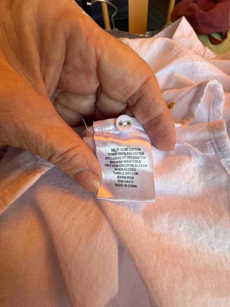 May include: Close-up of a white garment with a fabric tag. The tag reads "SELF: 100% COTTON, TRIMS: 100% POLYESTER, EXCLUSIVE OF DECORATION, MACHINE WASH COLD, ONLY NON-CHLORINE BLEACH WHEN NEEDED, TUMBLE DRY LOW, WARM IRON, RN#149414, MADE IN CHINA."