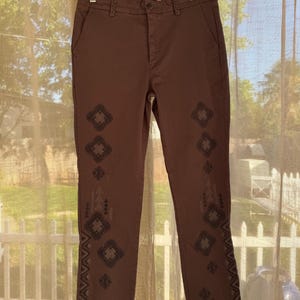 May include: Brown trousers with decorative geometric patterns down the sides. The trousers are hanging on a black hanger. The trousers have a button and zip closure. The trousers are made of a sturdy material.