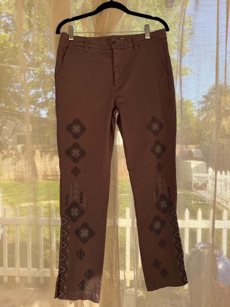 May include: Brown trousers with decorative geometric patterns down the sides. The trousers are hanging on a black hanger. The trousers have a button and zip closure. The trousers are made of a sturdy material.