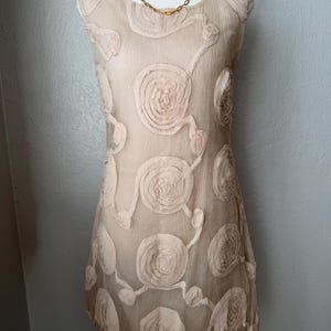 May include: A beige sleeveless dress with a floral design. The dress features a round neckline and is adorned with raised, circular floral appliques. A gold-colored layered necklace is draped on the mannequin. The dress is displayed on a white mannequin.