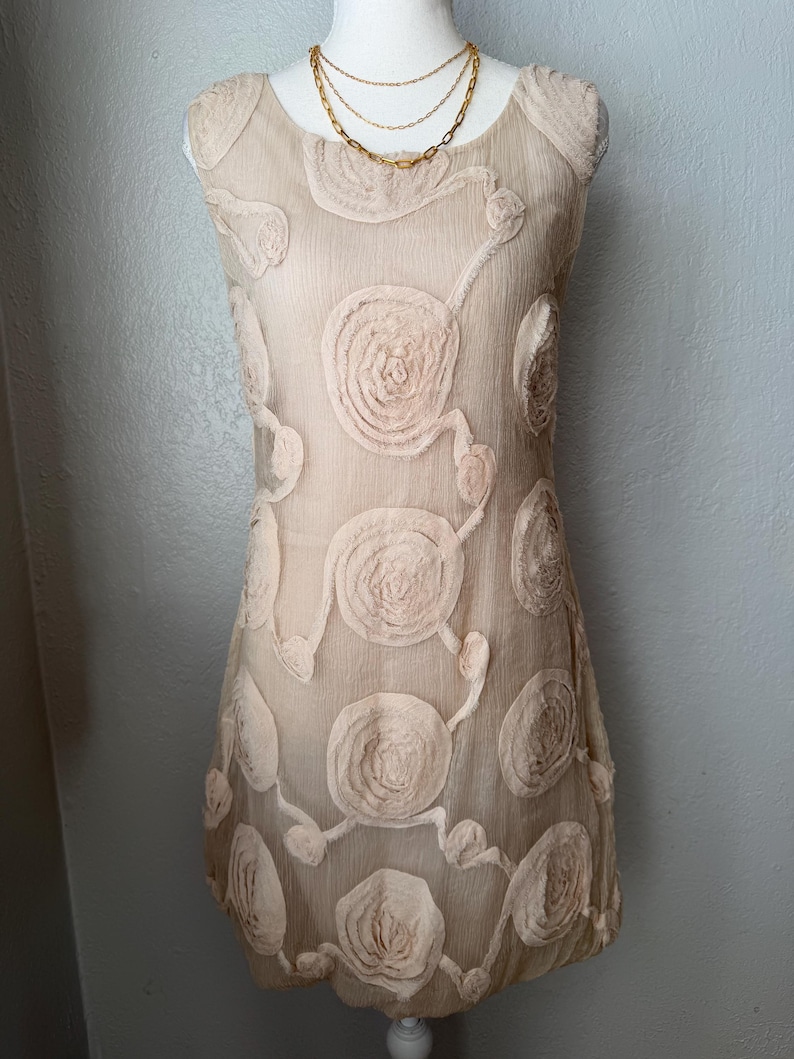 May include: A beige sleeveless dress with a floral design. The dress features a round neckline and is adorned with raised, circular floral appliques. A gold-colored layered necklace is draped on the mannequin. The dress is displayed on a white mannequin.