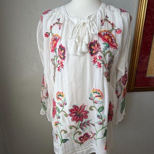 May include: White tunic top with colorful floral embroidery. The long-sleeved top features red, pink, yellow, and green embroidered flowers and leaves. It has a round neckline with a tie and tassel detail.